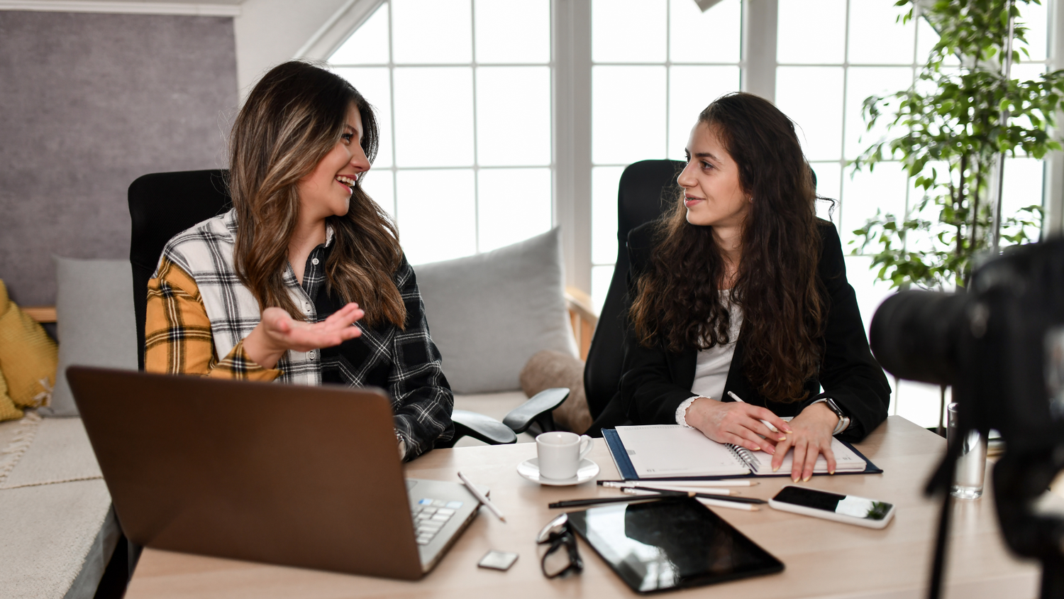 Two women recording a video podcast interview at a table with a laptop, notebook, and a camera capturing them