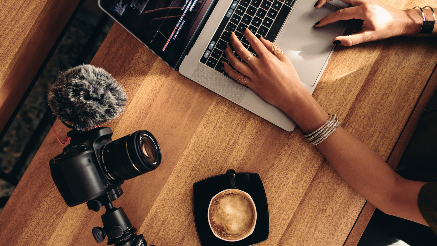 Overhead view of hands typing on a laptop running a video editor, next to a camera fitted with a windscreen microphone and a cup of coffee