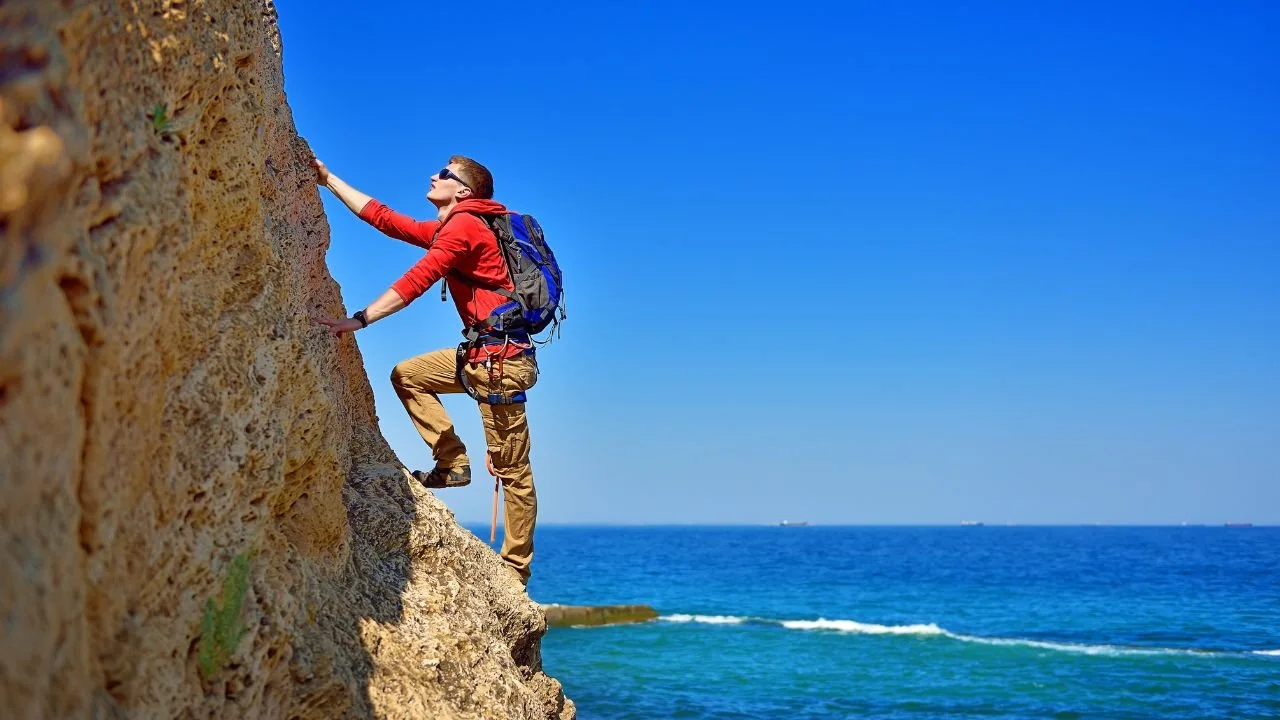 A rock climber in a red jacket scaling a steep cliff face high above the ocean