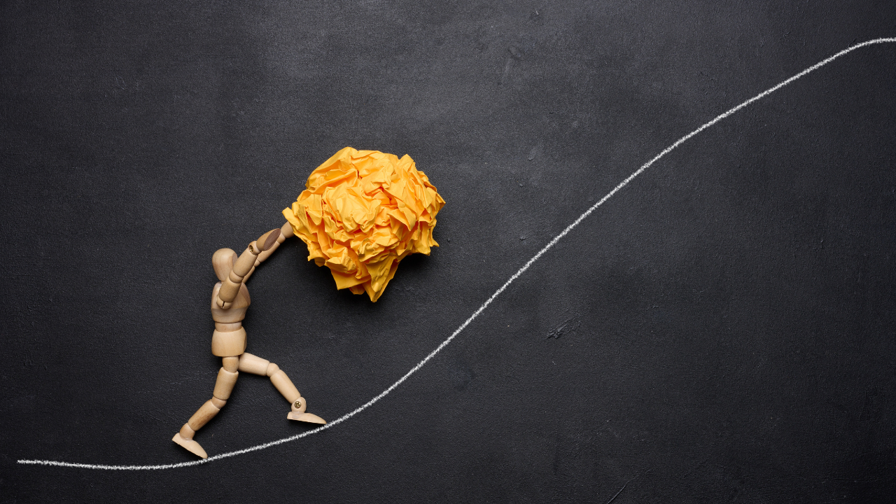A wooden artist's mannequin pushing a large crumpled paper ball up a hill drawn in chalk, symbolizing effort against the odds