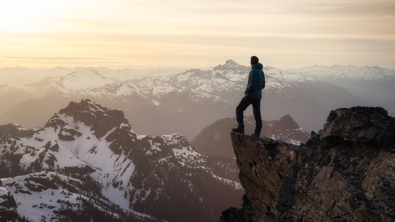 A lone figure standing at the edge of a rocky mountain peak at sunrise, overlooking a range of snow-capped mountains